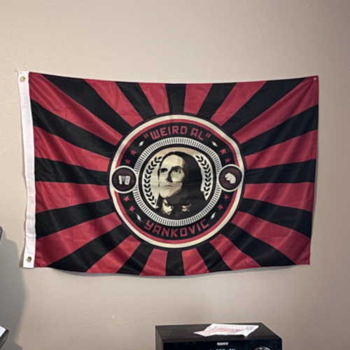 Photograph of a flag based on the imagery of Mandatory Fun, showing Al's face in the center, with a hamster on his left and an accordion on his right, with a black and red sunburst pattern behind them.